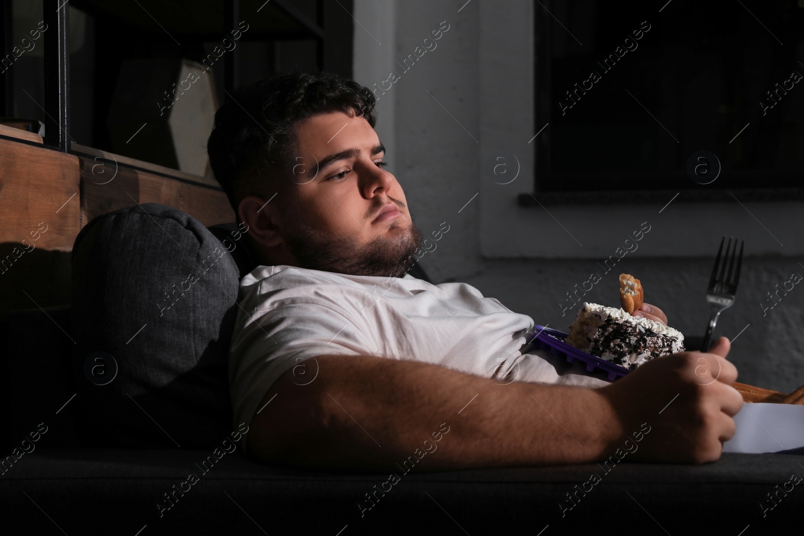 Photo of Depressed overweight man eating sweets in living room at night