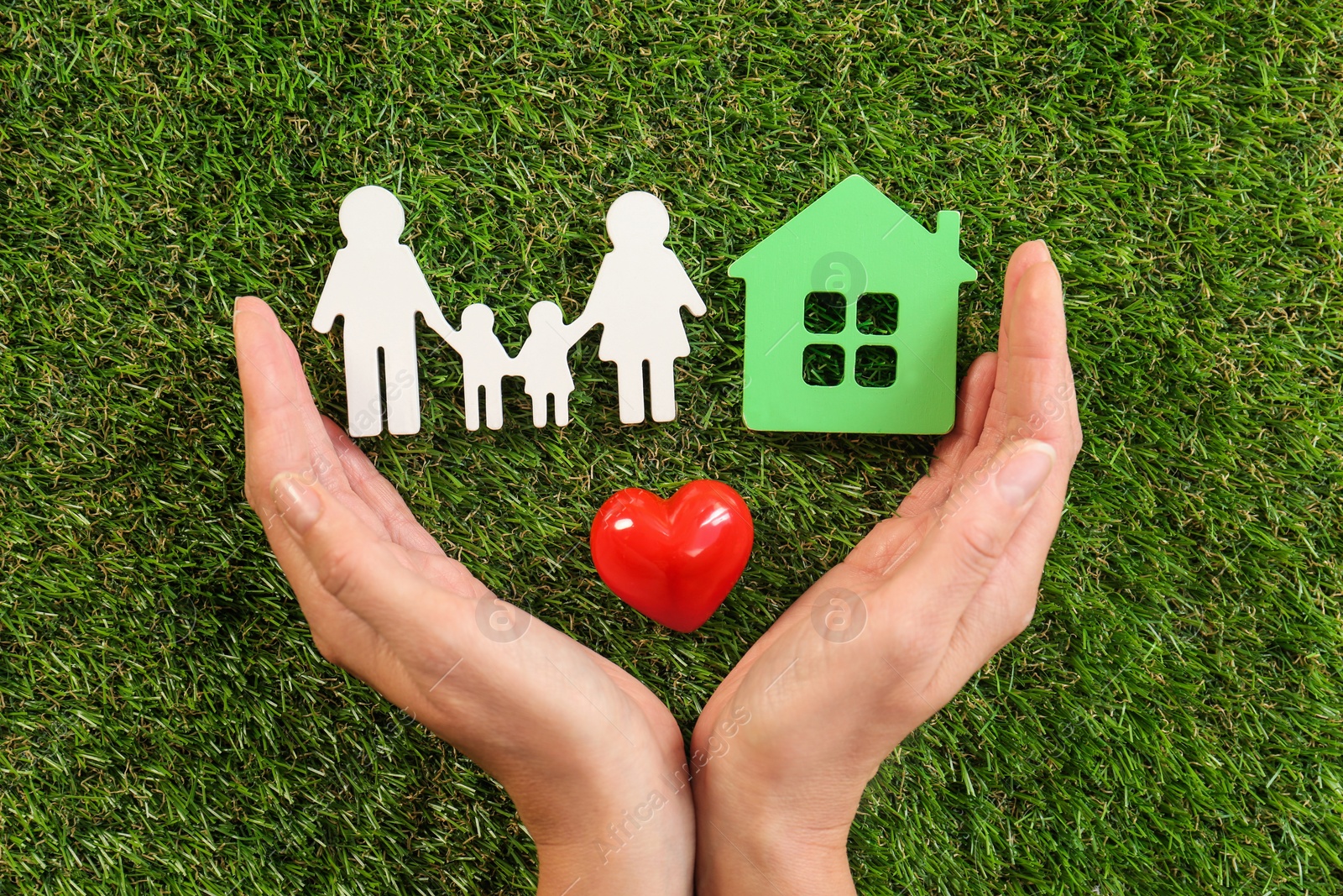 Photo of Woman holding hands near red heart and figures of house and family on green grass, top view