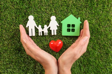 Photo of Woman holding hands near red heart and figures of house and family on green grass, top view