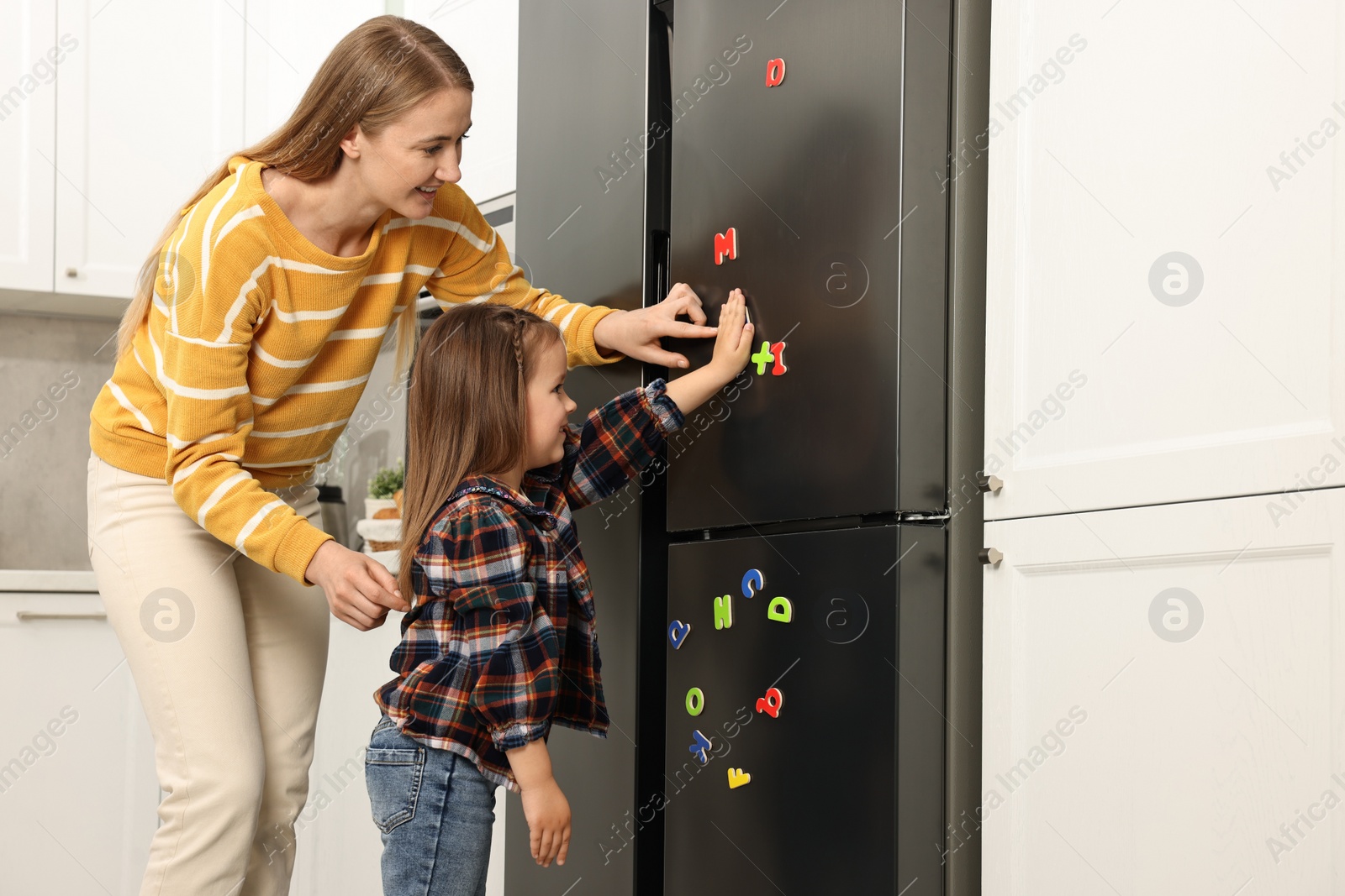 Photo of Mom and daughter putting magnetic letters on fridge at home. Learning alphabet