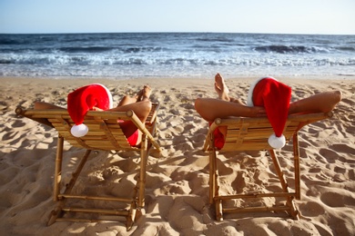 Photo of Lovely couple with Santa hats relaxing on deck chairs at beach, back view. Christmas vacation