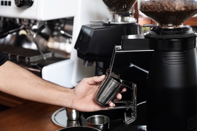 Photo of Barista with metal cup using coffee grinding machine in cafe, closeup