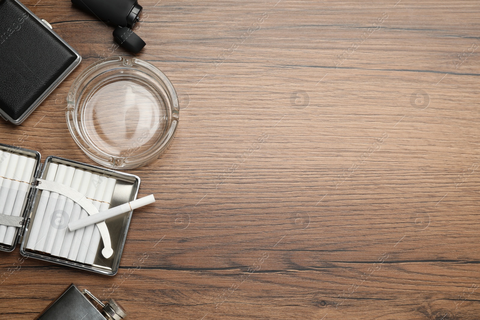 Photo of Open case with tobacco filter cigarettes, glass ashtray, lighter and pocket flask on wooden table, flat lay. Space for text