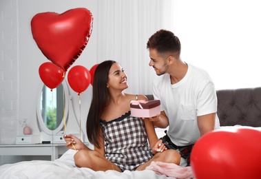 Photo of Young man presenting gift to his girlfriend in bedroom decorated with air balloons. Celebration of Saint Valentine's Day