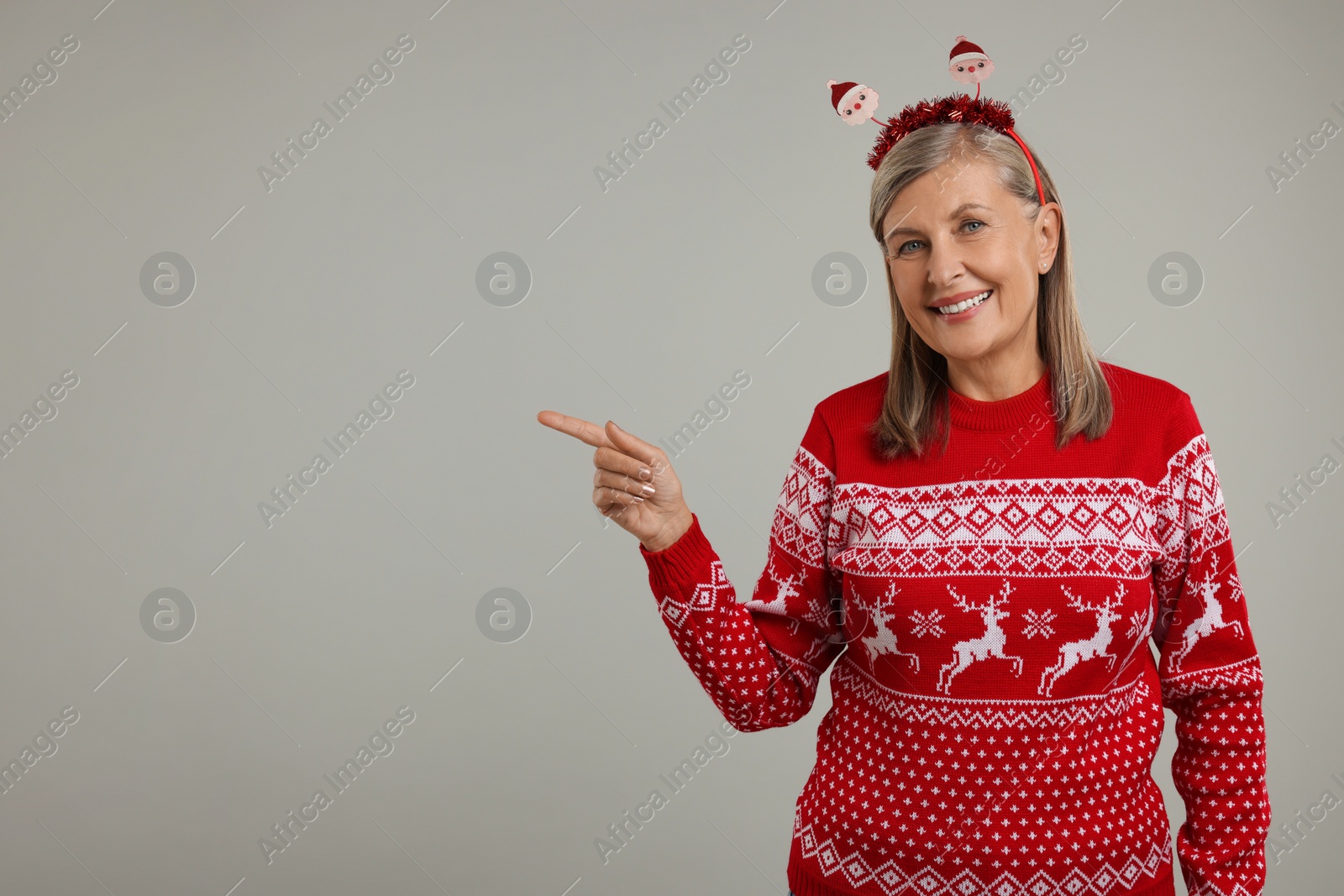 Photo of Happy senior woman in Christmas sweater and Santa headband pointing at something on grey background. Space for text