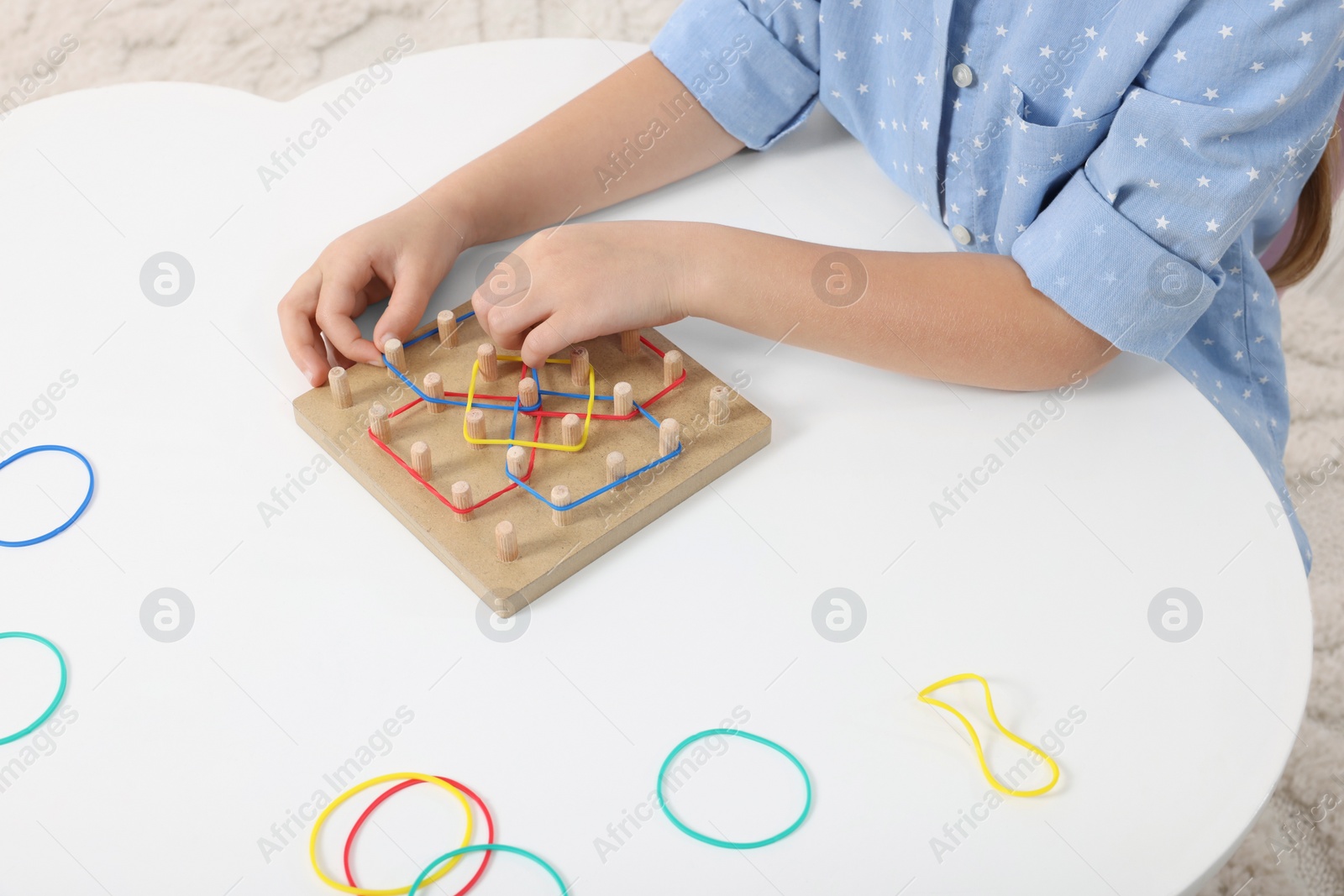 Photo of Motor skills development. Girl playing with geoboard and rubber bands at white table, closeup