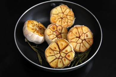 Photo of Frying pan with fried garlic and rosemary on black table, closeup