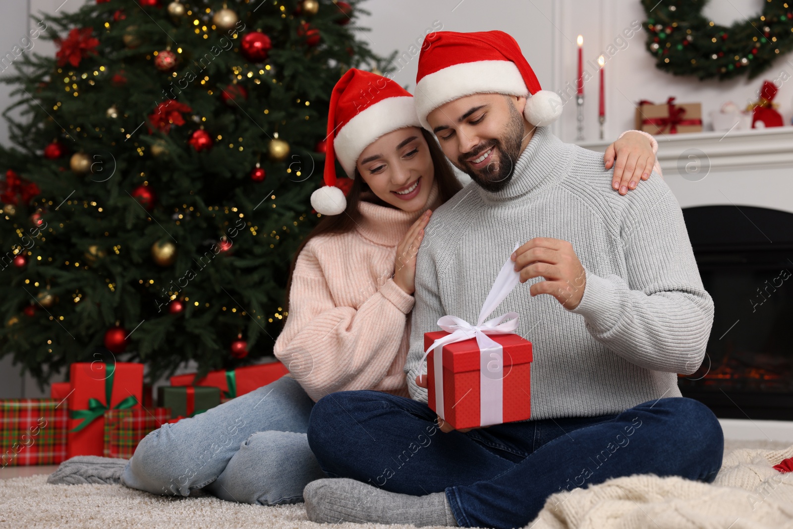 Photo of Happy woman in Santa hat presenting Christmas gift to her boyfriend at home