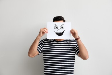 Photo of Man hiding emotions using card with drawn smiling face on white background