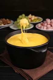 Dipping piece of ham into fondue pot with melted cheese on black wooden table, closeup