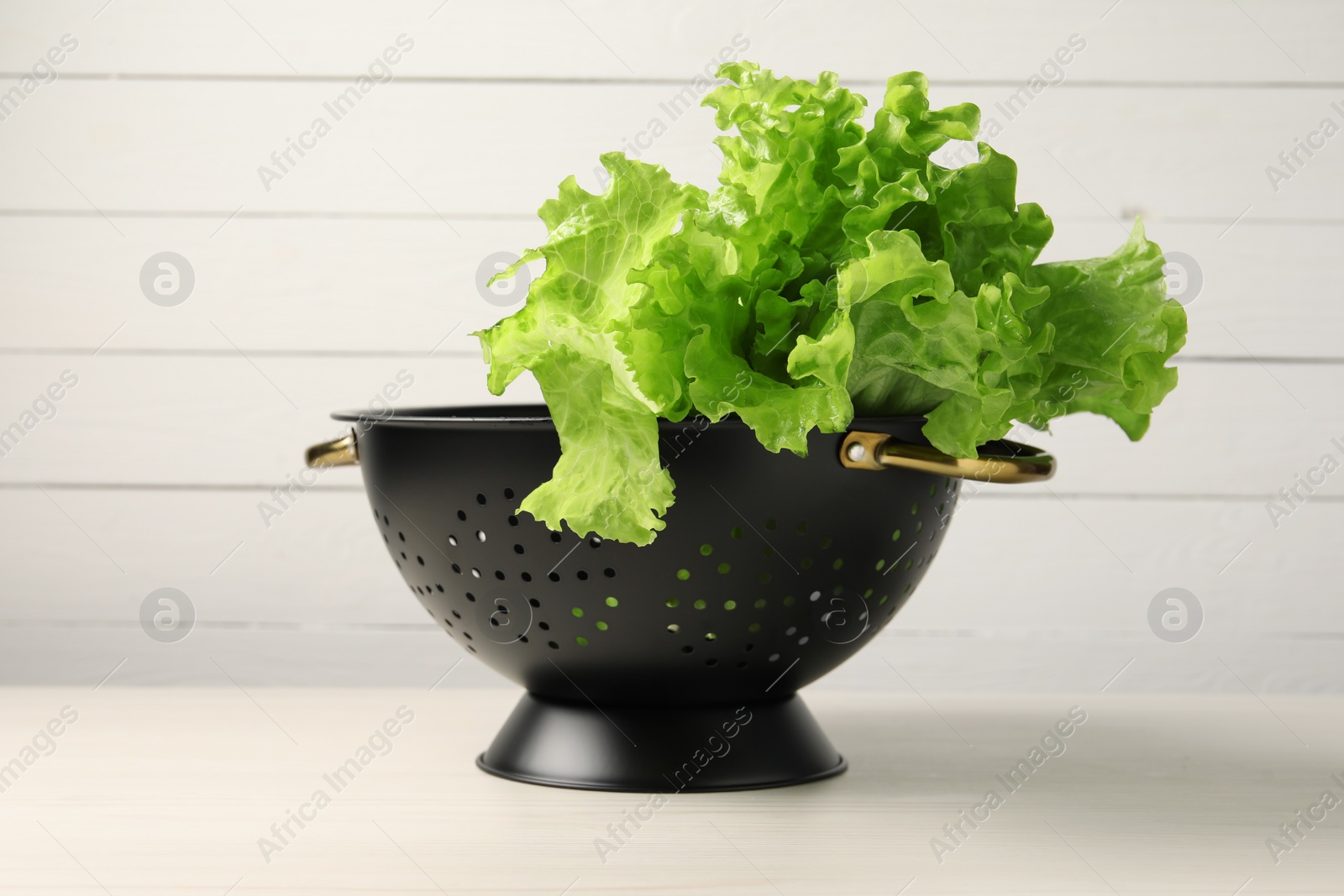 Photo of Fresh lettuce in black colander on white wooden table