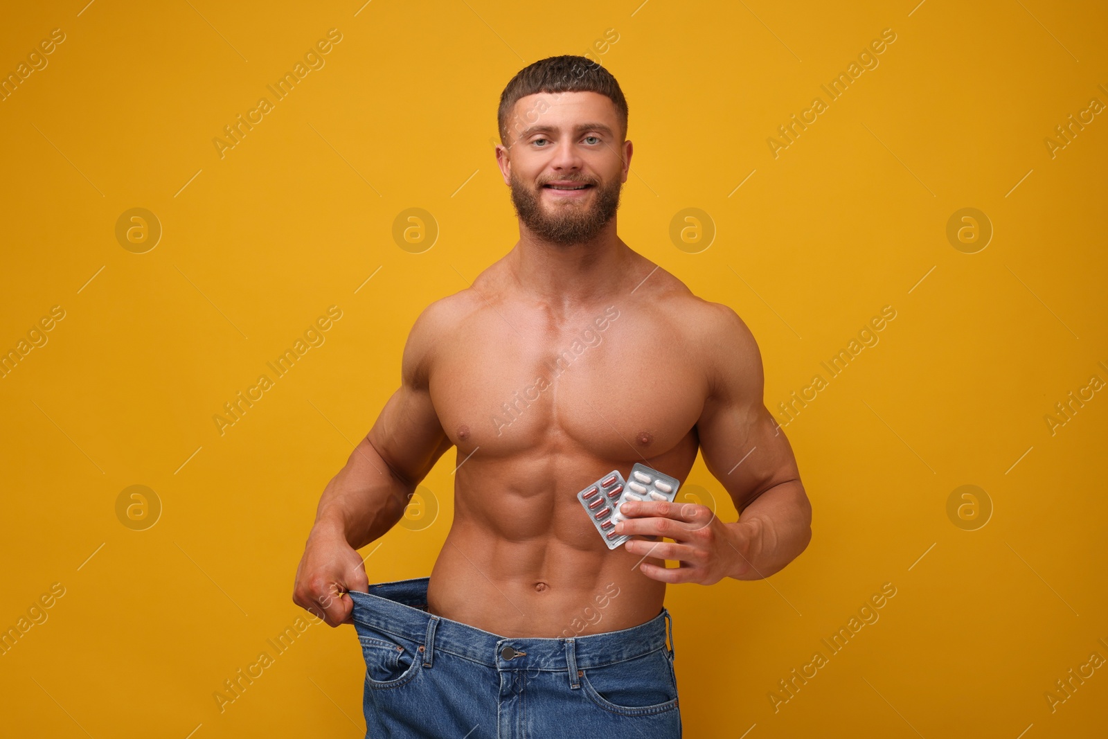 Photo of Athletic young man with pills wearing big jeans on orange background. Weight loss