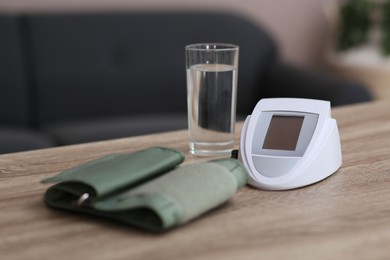 Modern blood pressure monitor and glass of water on wooden table indoors, closeup