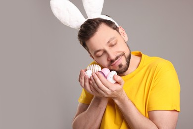 Photo of Happy man in bunny ears headband holding painted Easter eggs on grey background. Space for text