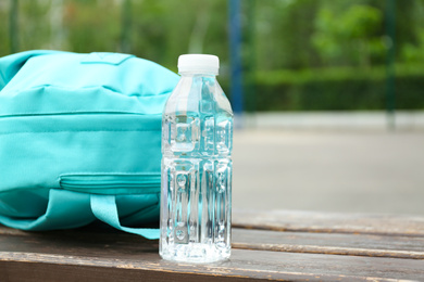 Photo of Bottle with water and turquoise backpack on wooden bench outdoors, closeup. Space for text