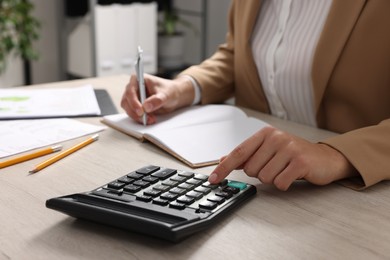 Woman using calculator while taking notes at wooden table indoors, closeup