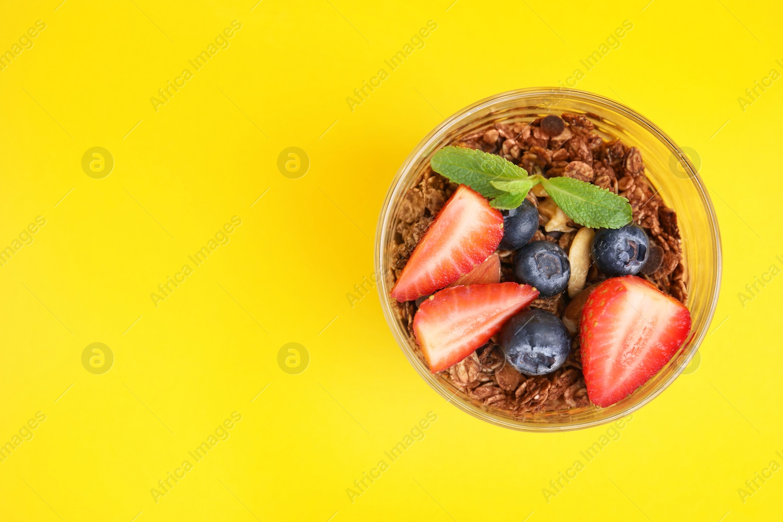 Photo of Tasty granola with berries and mint in glass on yellow background, top view. Space for text