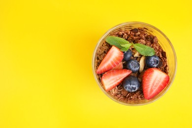 Photo of Tasty granola with berries and mint in glass on yellow background, top view. Space for text