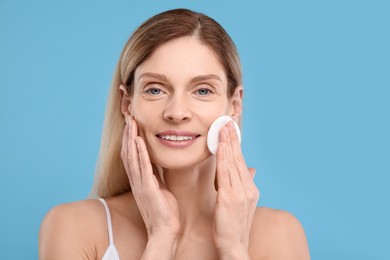 Beautiful woman removing makeup with cotton pad on light blue background