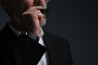 Photo of Bearded man smoking cigar against dark grey background, closeup. Space for text