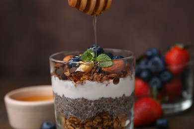 Photo of Pouring honey onto tasty granola with berries, yogurt and chia seeds at table, closeup