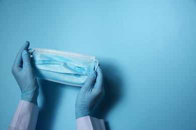 Photo of Doctor in latex gloves holding disposable face mask on light blue background, closeup with space for text. Protective measures during coronavirus quarantine
