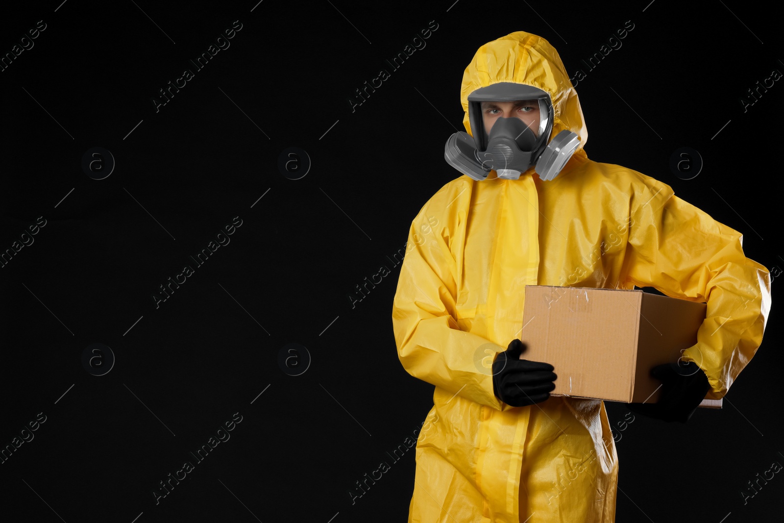 Photo of Man wearing chemical protective suit with cardboard box on black background, space for text. Prevention of virus spread