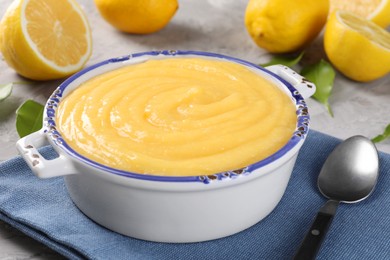 Delicious lemon curd in bowl, fresh citrus fruits, green leaves and spoon on grey table, closeup