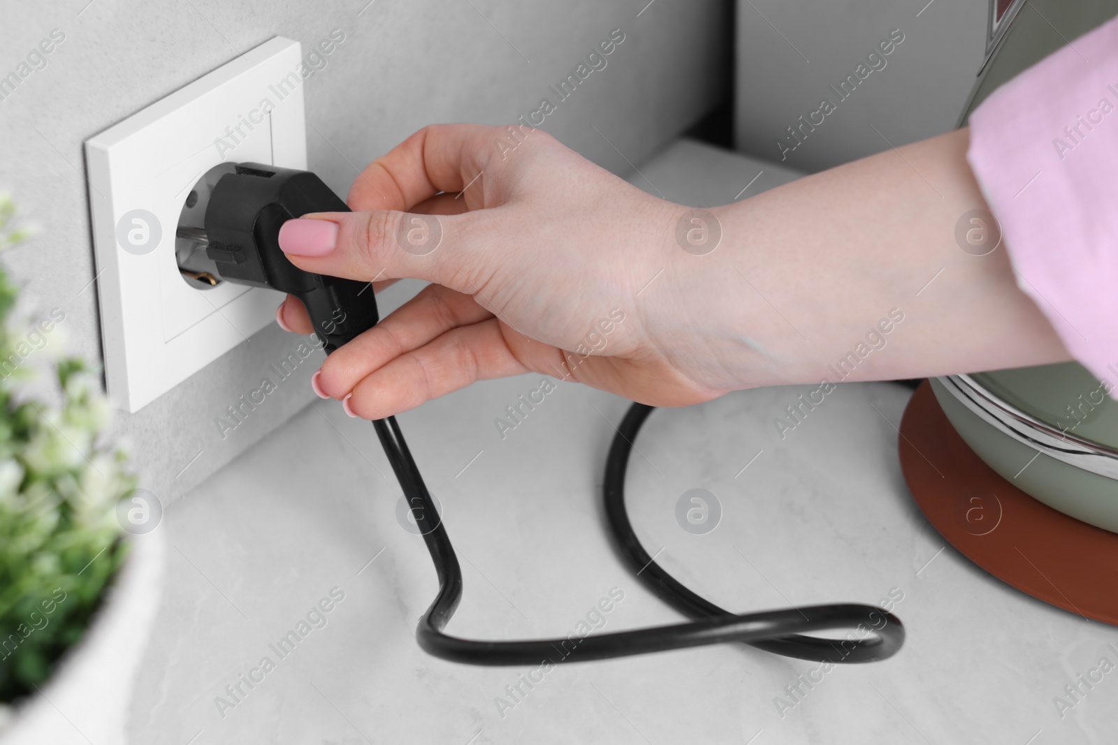 Photo of Woman plugging electric kettle into power socket at white table indoors, closeup