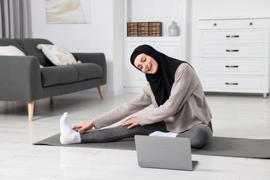 Photo of Muslim woman in hijab stretching near laptop on fitness mat at home