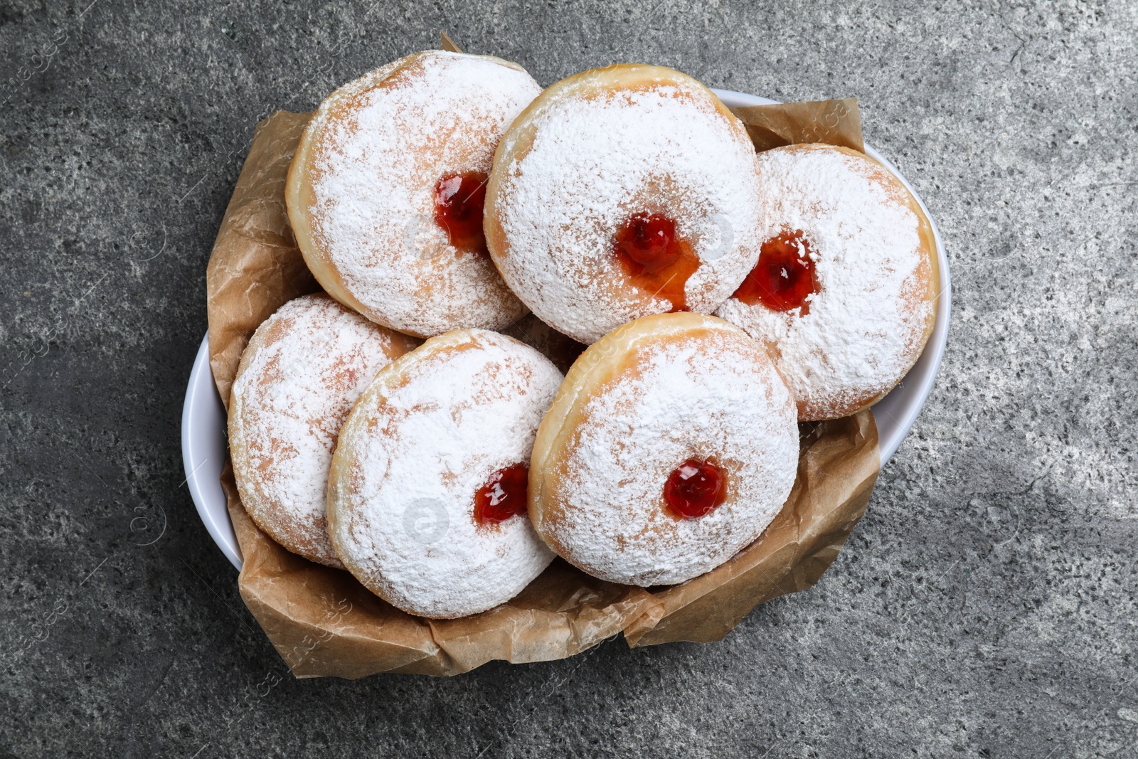 Photo of Delicious donuts with jelly and powdered sugar in bowl on grey table, top view