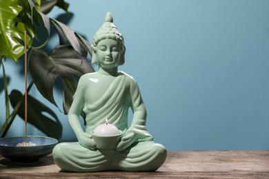 Photo of Buddhism religion. Decorative Buddha statue with burning candle, incense stick on wooden table and monstera against light blue wall, space for text
