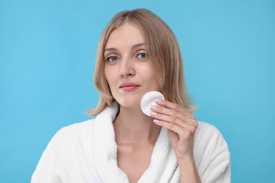 Young woman cleaning face with cotton pad on light blue background