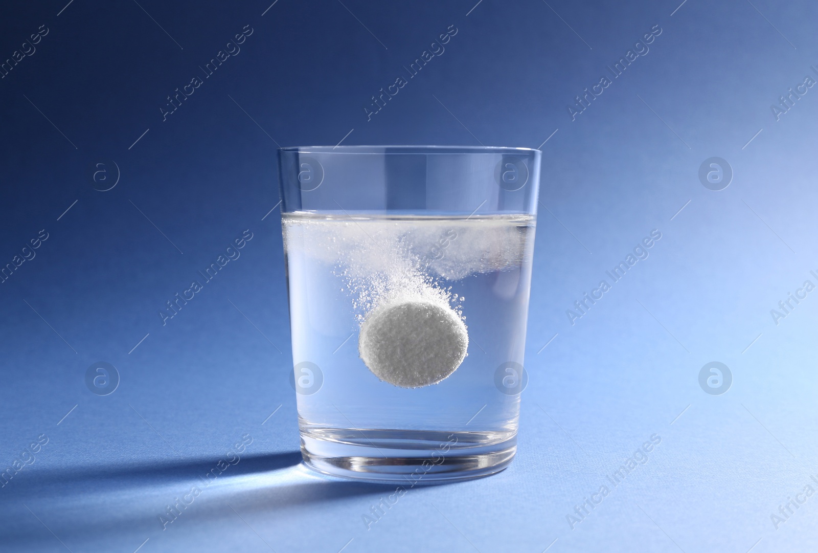 Photo of Effervescent pill dissolving in glass of water on blue background