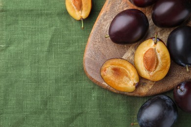 Many tasty ripe plums and wooden board on green fabric, flat lay. Space for text