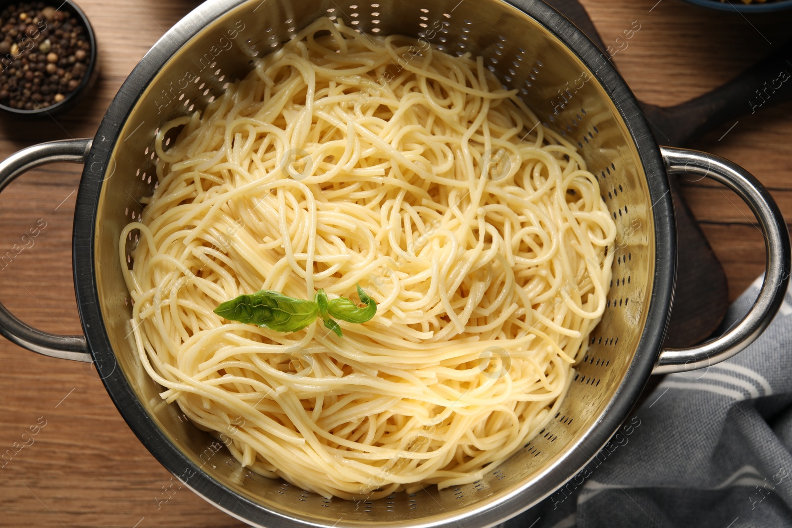 Photo of Cooked pasta in metal colander and peppercorns on wooden table, top view