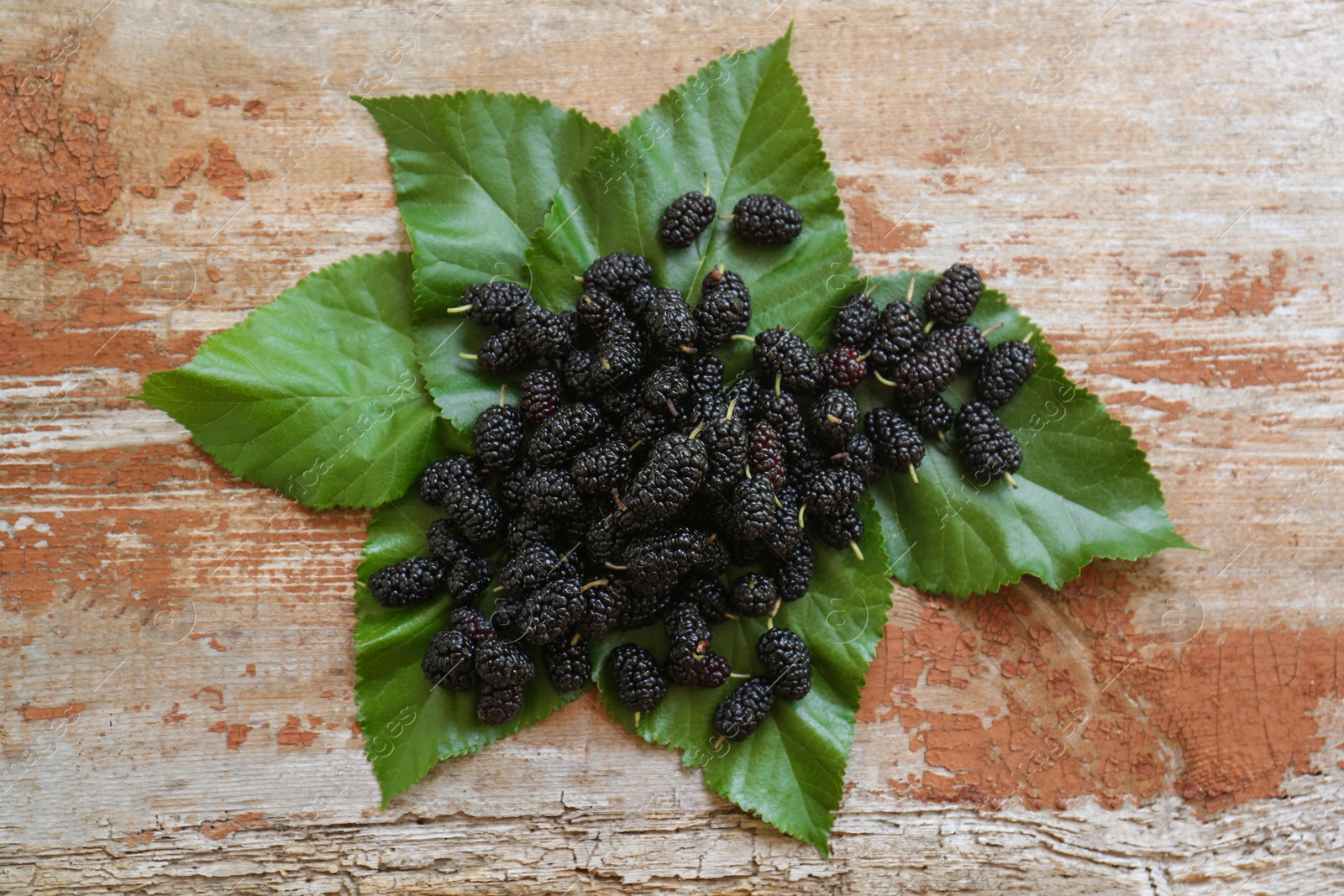 Photo of Heap of delicious ripe black mulberries and green leaves on wooden table, flat lay