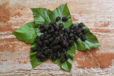 Heap of delicious ripe black mulberries and green leaves on wooden table, flat lay