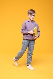 Photo of Cute schoolboy in glasses holding books and walking on orange background