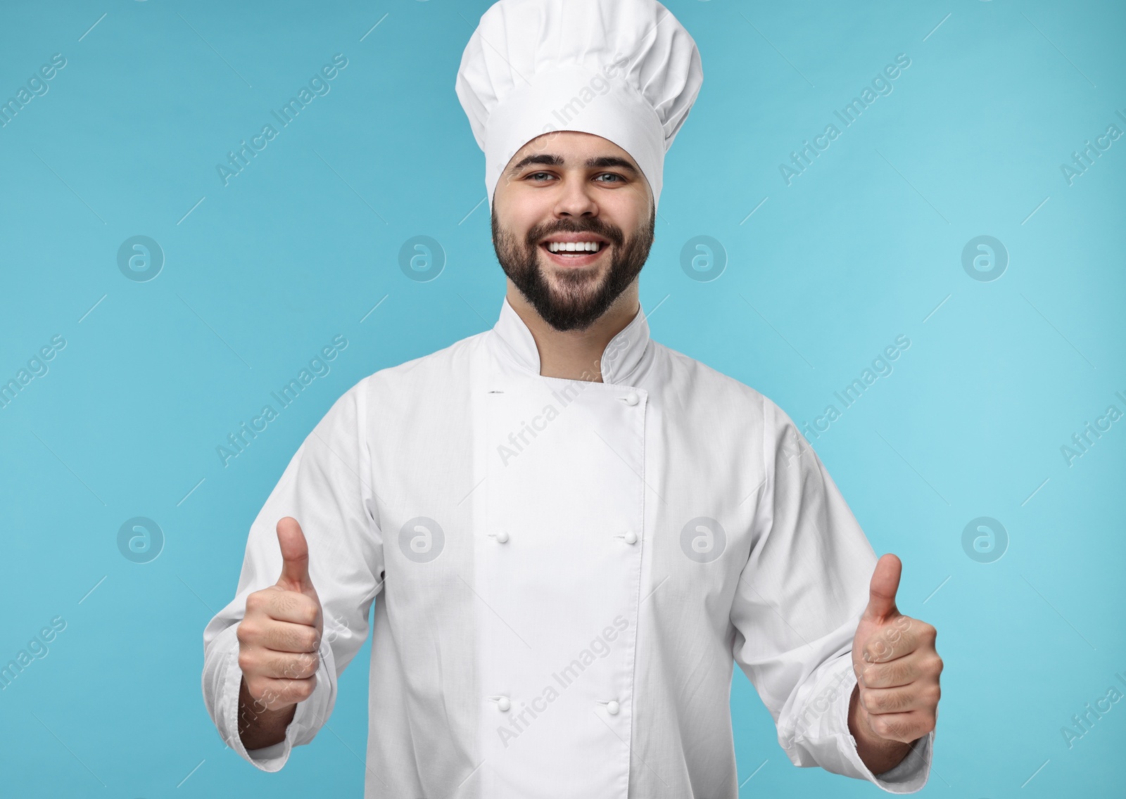 Photo of Happy young chef in uniform showing thumbs up on light blue background