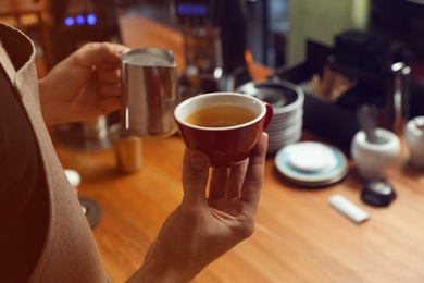 Photo of Barista holding cup of coffee and jug with milk at bar counter, closeup. Space for text