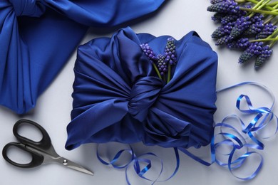 Furoshiki technique. Gift packed in blue silk fabric, muscari flowers, ribbon and scissors on white table, flat lay