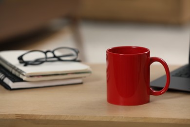 Red ceramic mug, notebooks and laptop on wooden table at workplace. Space for text