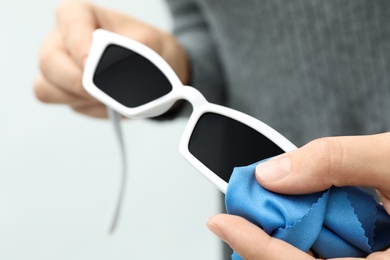 Woman wiping sunglasses with microfiber cleaning cloth on light background, closeup