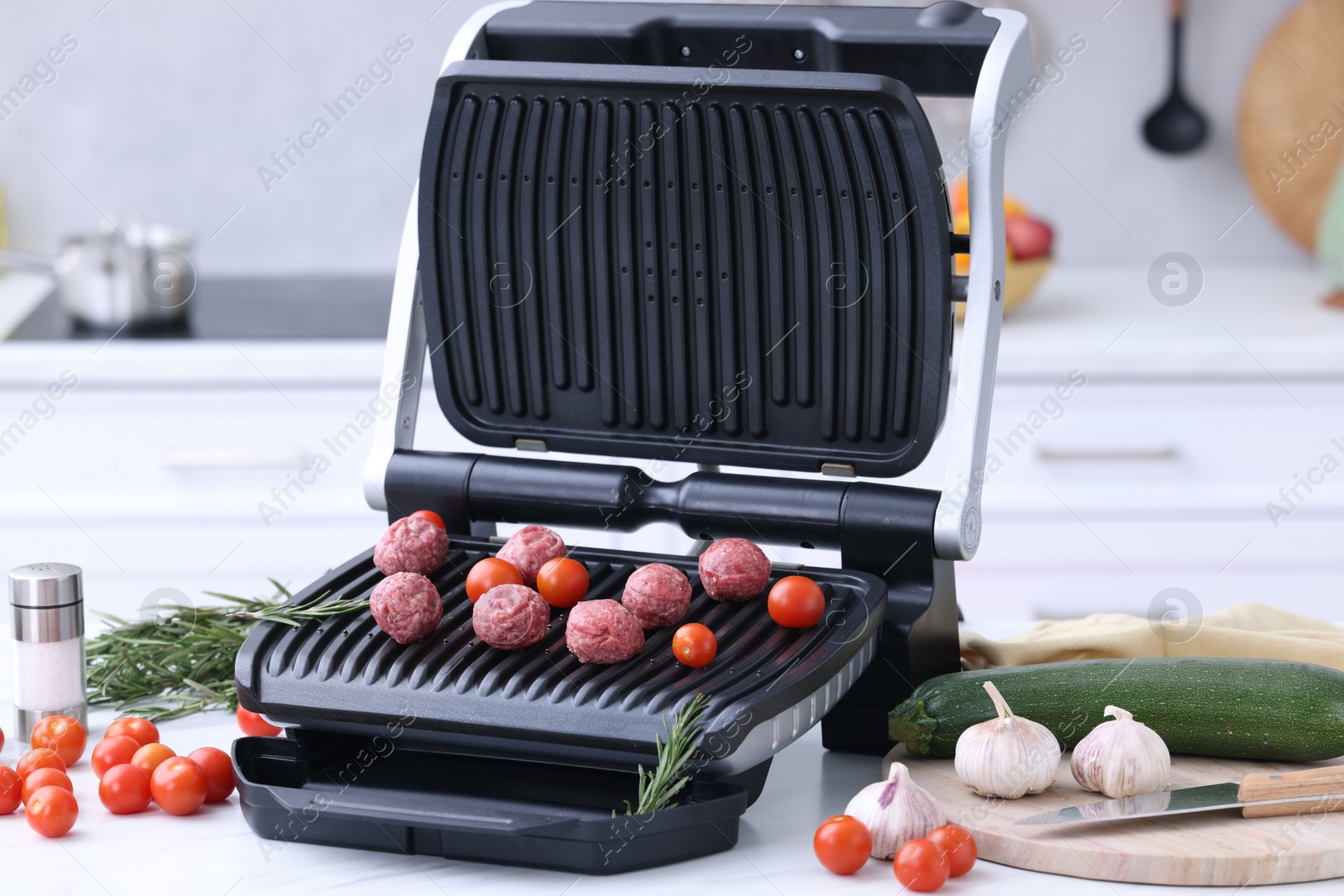 Photo of Electric grill with meatballs, tomatoes and vegetables on white table in kitchen