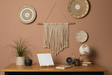 Photo of Stylish macrame and wicker wall decor hanging above wooden table with houseplants and stationery