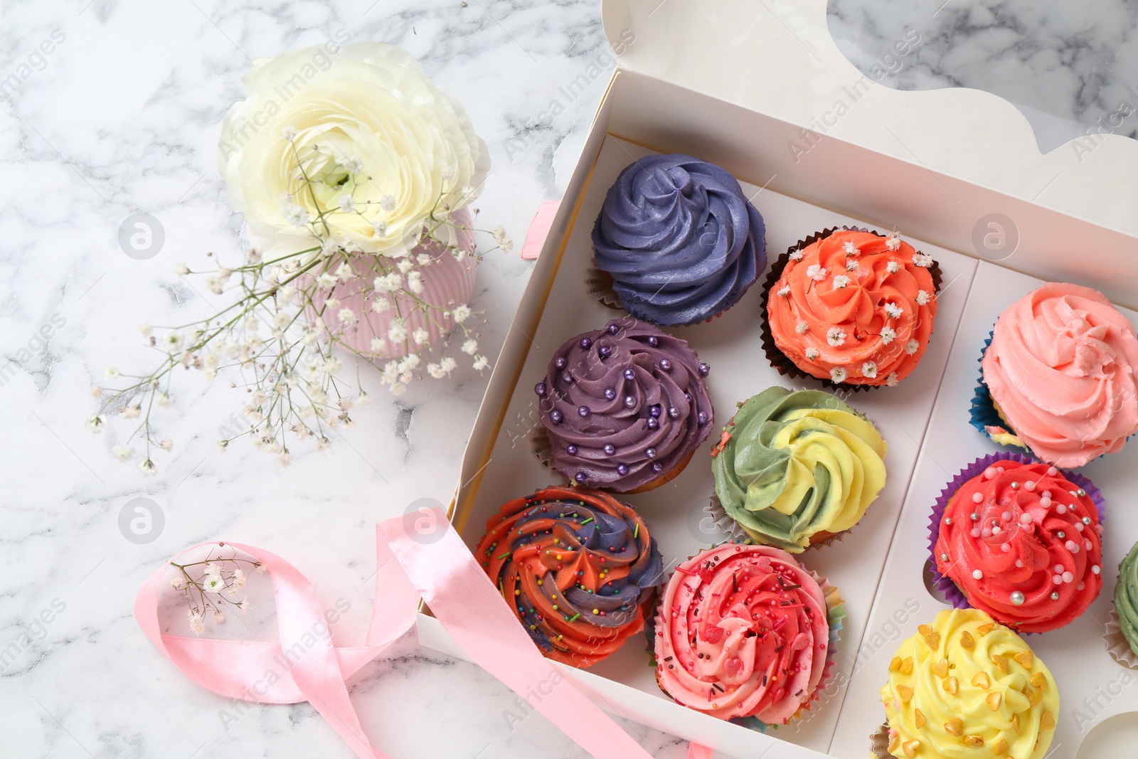 Photo of Different colorful cupcakes in box and flowers on white marble table, top view