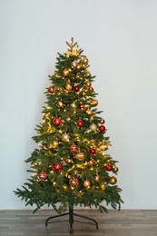 Beautiful Christmas tree with fairy lights and festive decor near white wall