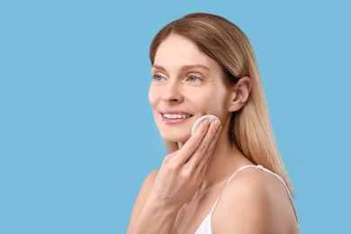 Beautiful woman removing makeup with cotton pad on light blue background, space for text