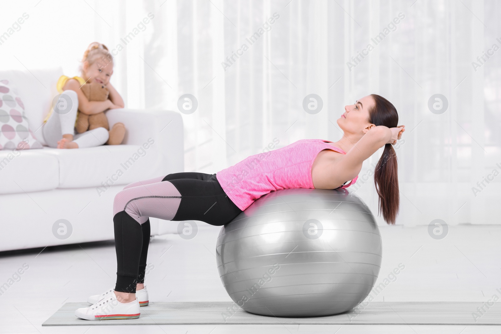 Photo of Woman doing fitness exercises while her daughter sitting on sofa at home